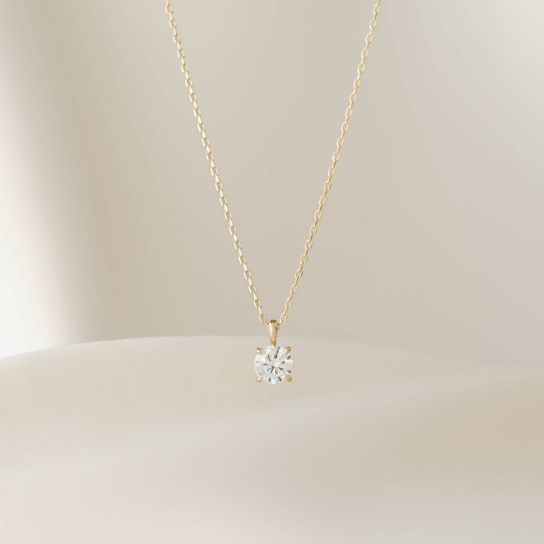 A close-up shot of a delicate Gold necklace with a small, round diamond solitaire pendant in a prong setting, draped over a textured, light beige surface.