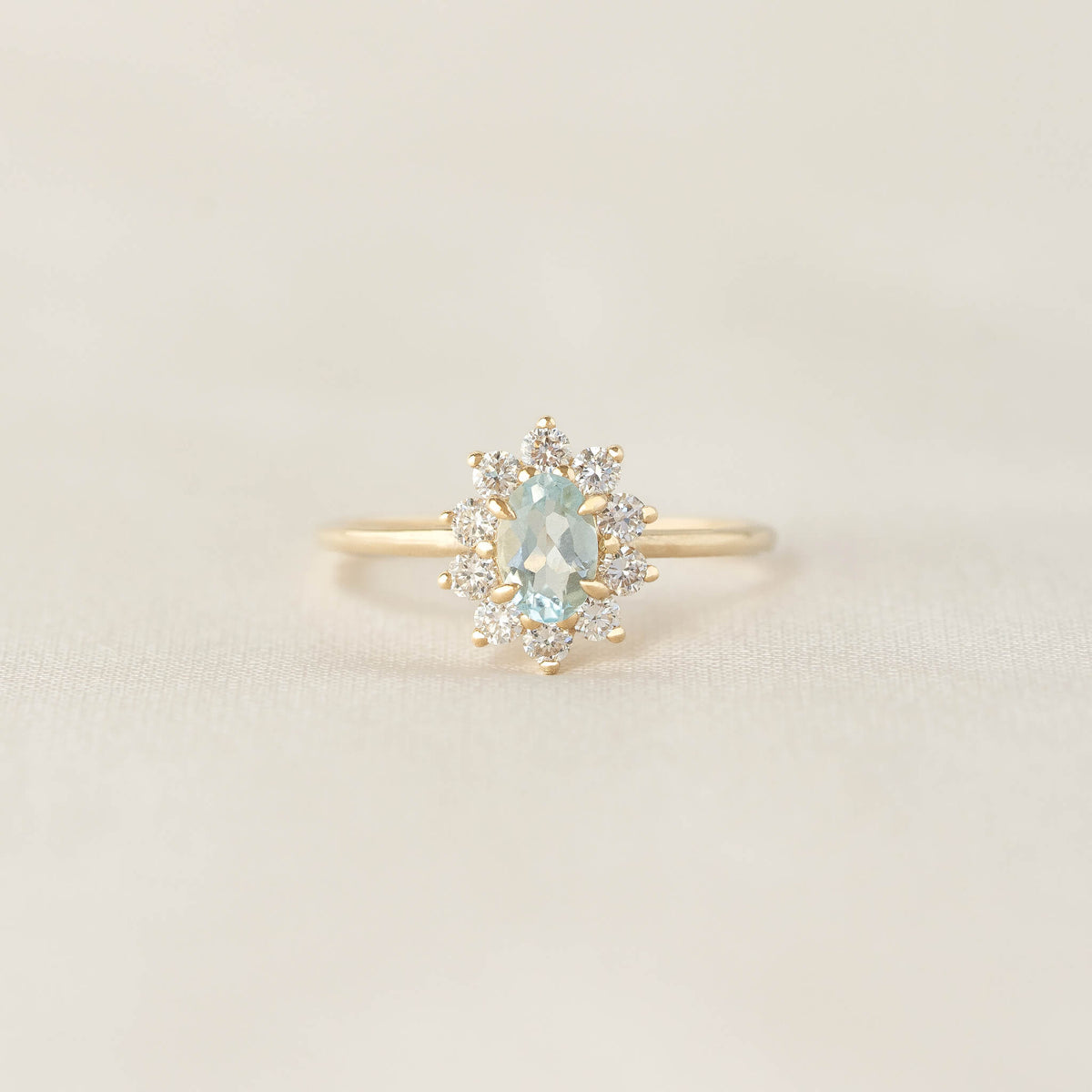 A delicate gold ring features a teal oval gemstone, encircled by small sparkling diamonds, set against a soft beige background, exuding elegance.