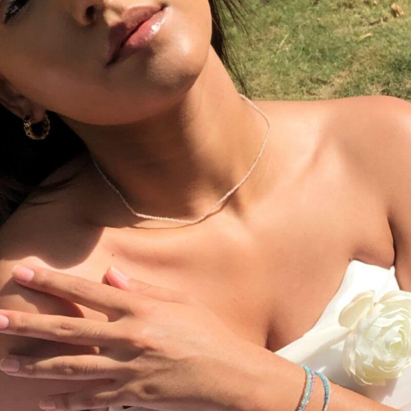 Close-up of a woman outdoors wearing a thin silver necklace, gold hoop earrings, and bracelets with a white floral detail on her top