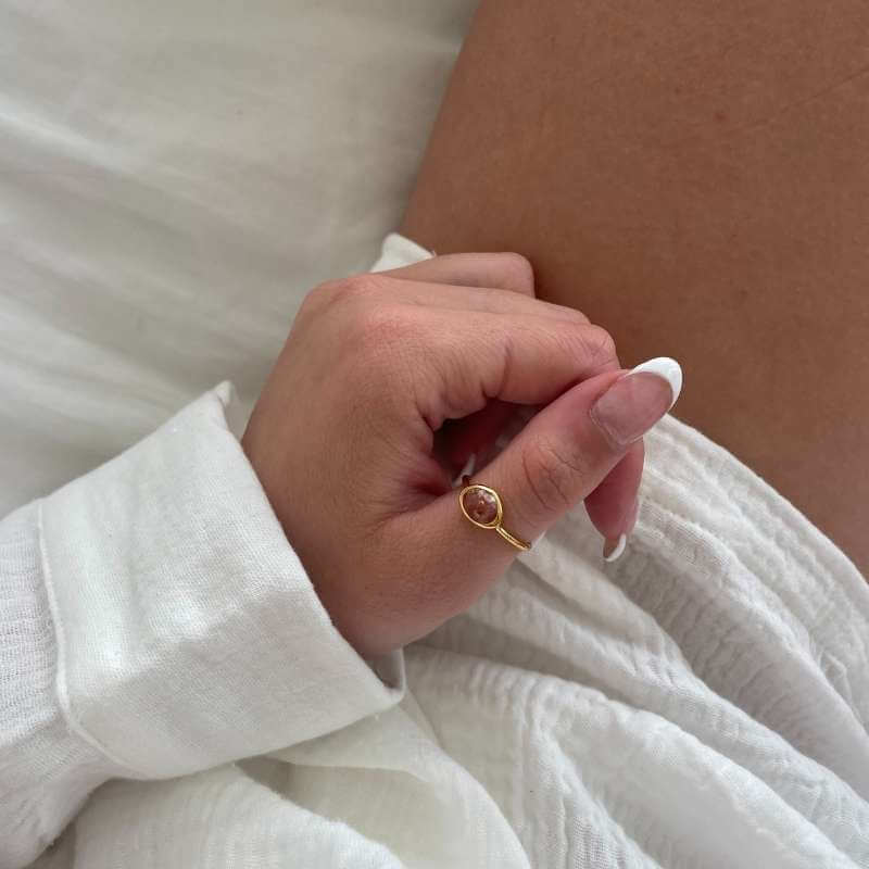 Close-up of a hand with natural nails wearing a gold ring with a brown stone resting on white fabric and skin background