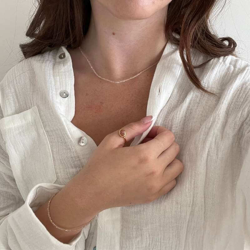 Close-up of a woman wearing a delicate gold ring, a thin necklace, and a matching bracelet with a white textured shirt