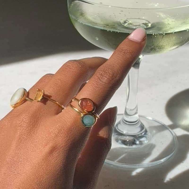 Close-up of a hand wearing four gold rings with colorful stones holding the stem of a glass with a light beverage