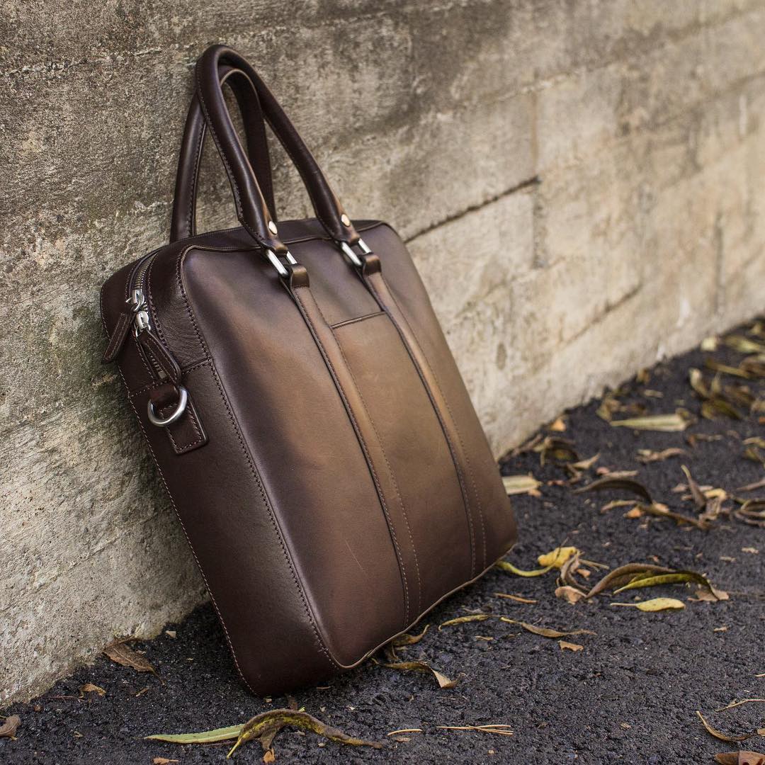 Close-up of a brown leather briefcase resting against a textured concrete wall with scattered dry leaves nearby