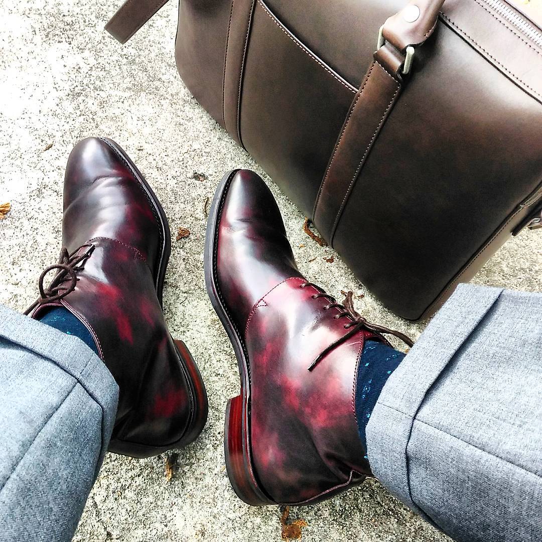 Close-up of burgundy leather dress boots worn with gray trousers and blue patterned socks on a concrete surface