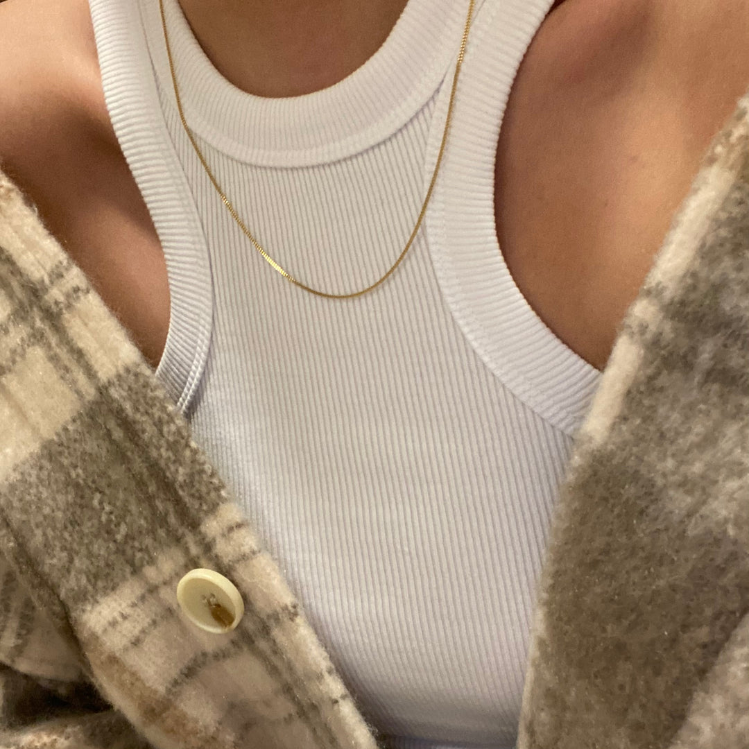 Close-up of a person wearing a white ribbed tank top, thin gold necklace, and a beige plaid wool jacket with a button visible