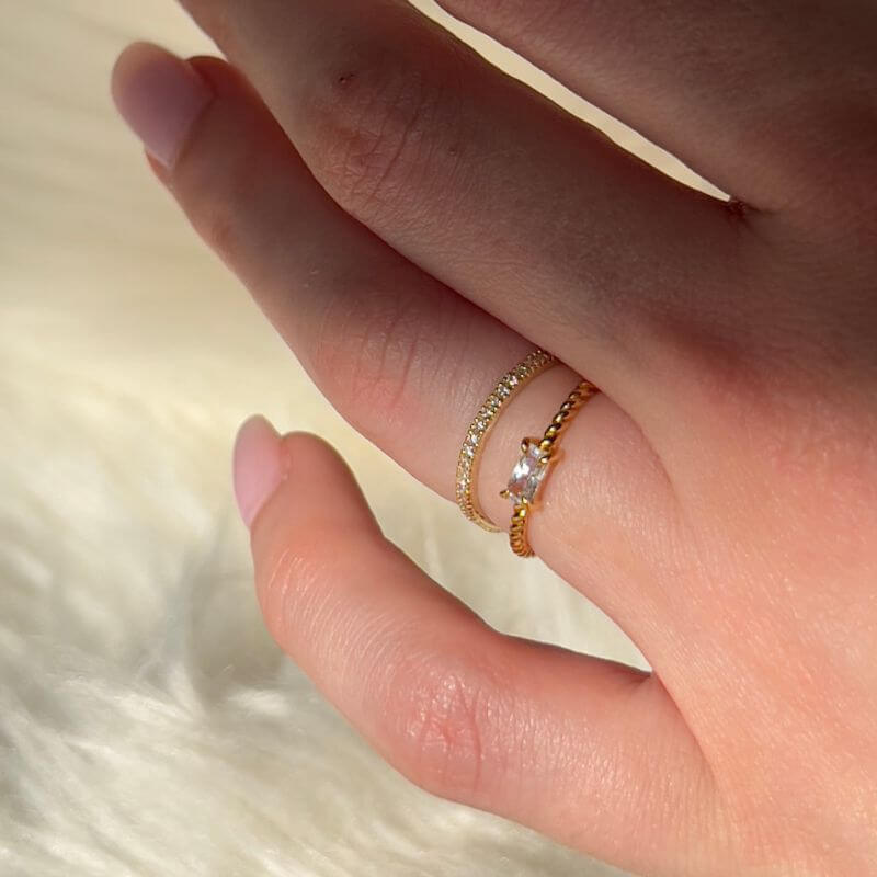 Close-up of a hand wearing two delicate gold rings, one with small clear stones and one with a single clear stone on a soft background
