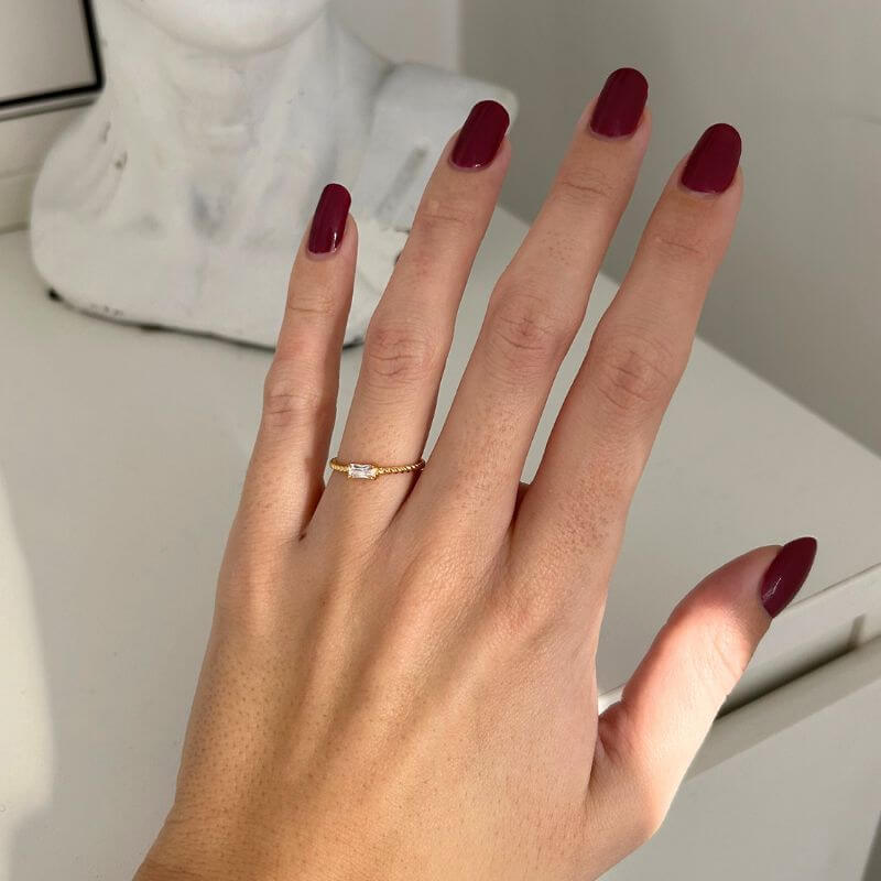 Close-up of a hand with dark red polished nails wearing a delicate gold ring with a small rectangular stone on a white surface
