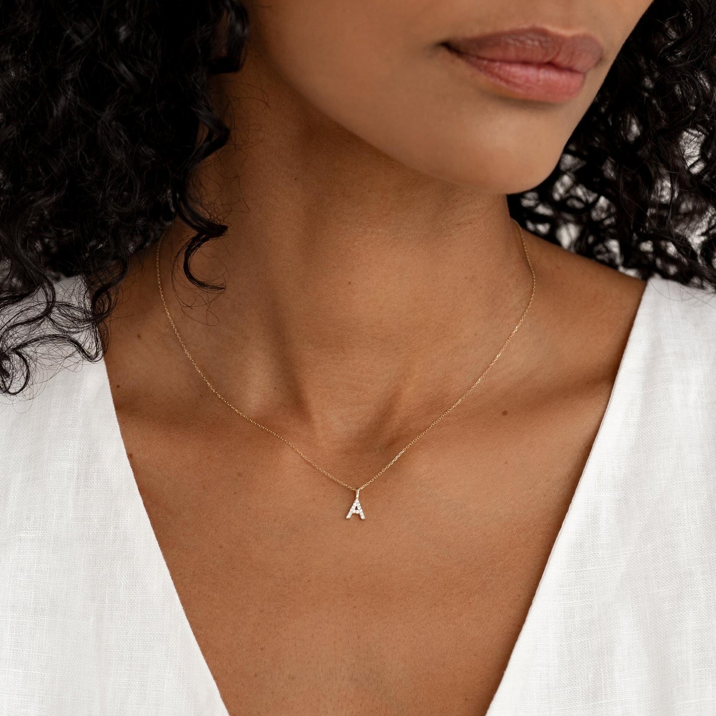 Close-up of a woman wearing a 14k gold cable chain necklace with a small letter A pendant and a white linen top