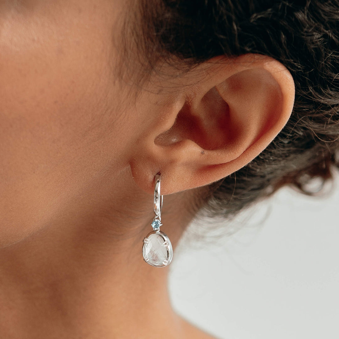 Close-up of a woman wearing Silver Rainbow Moonstone Earrings - Victoria (Blue Gem) with blue cubic zirconia accents