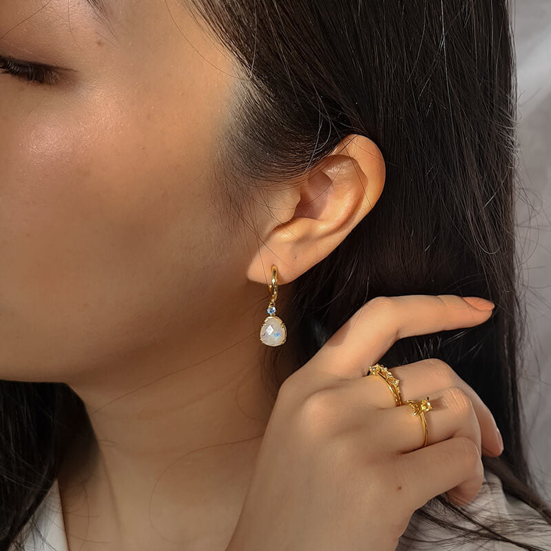 Close-up of a woman with dark hair wearing a dangling gold earring with a white stone and two gold rings on her hand