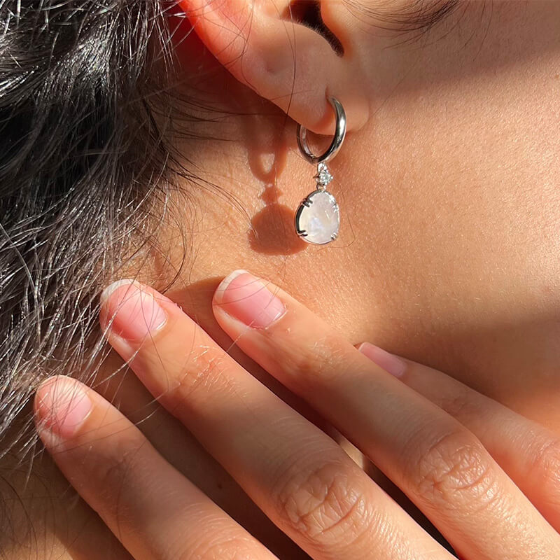 Close-up of an ear wearing a silver hoop earring with a teardrop-shaped clear gemstone pendant and fingers touching the neck