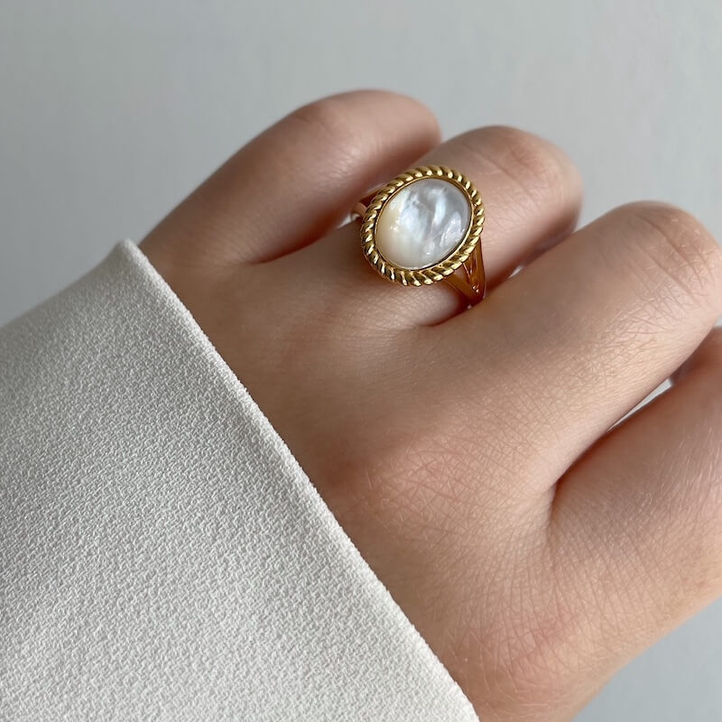 Close-up of a hand wearing a gold ring with an oval white mother-of-pearl centerpiece against a neutral background