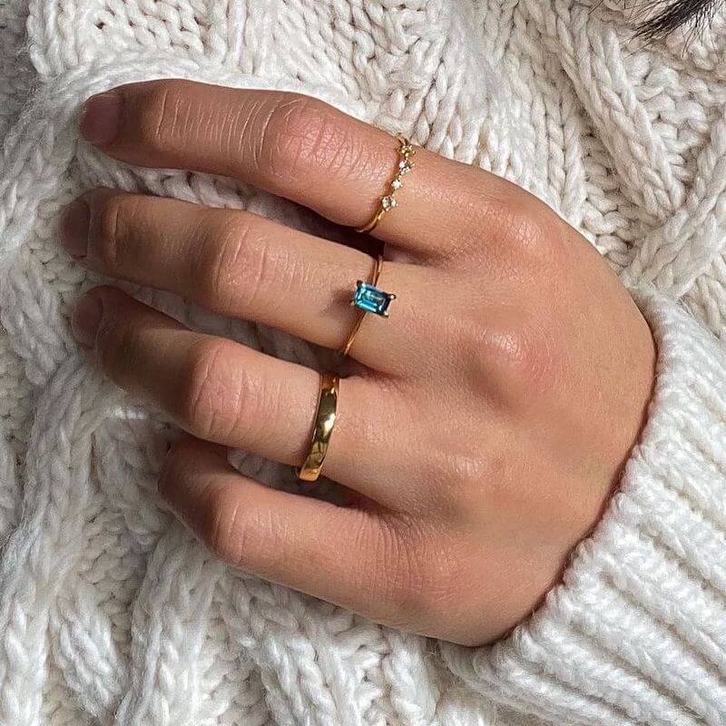 Close-up of a hand wearing three gold rings with a blue rectangular stone and small white gems on a white knitted sweater