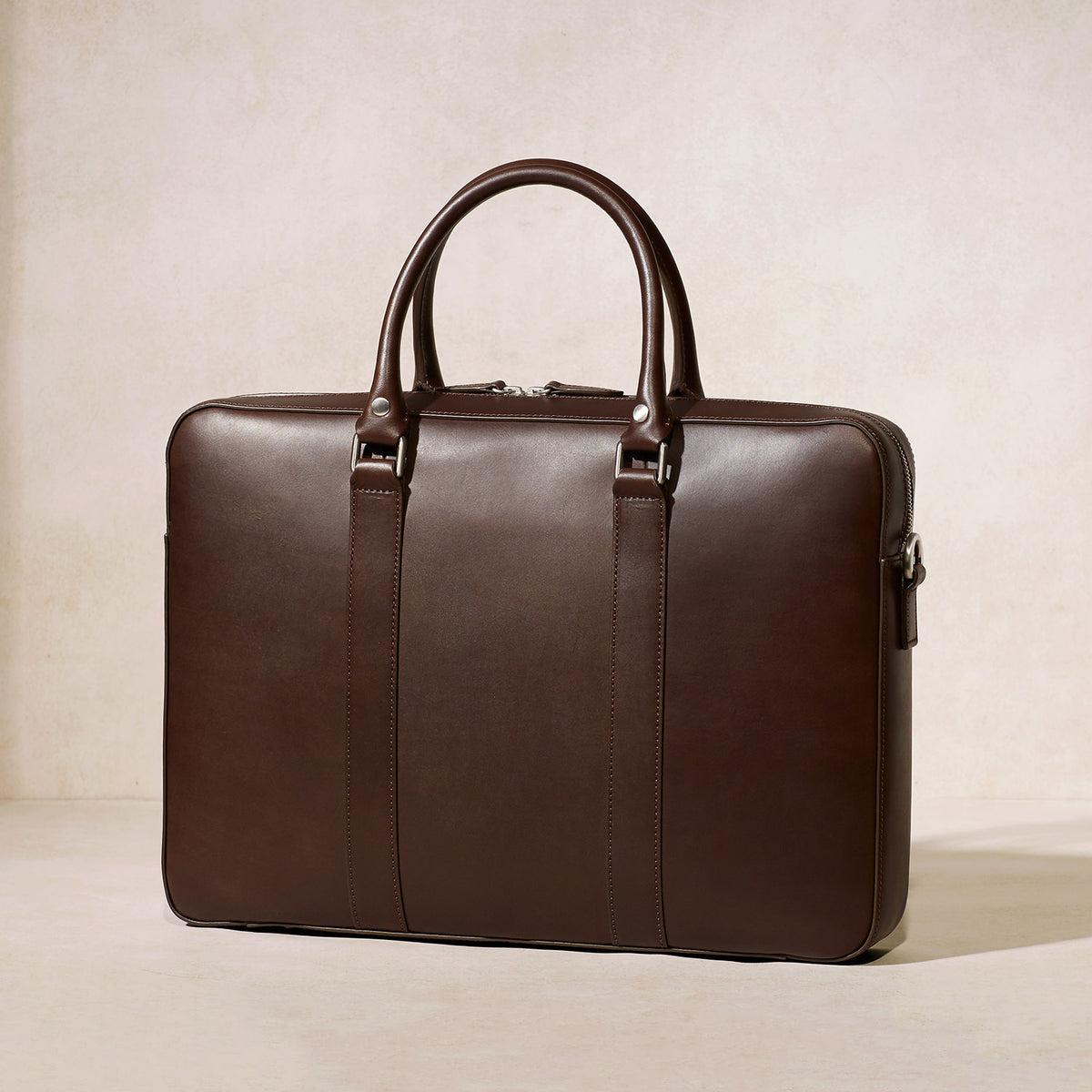 The Soft Briefcase (Private) in smooth dark brown leather with a slim and refined silhouette on a neutral background