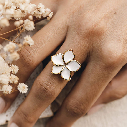 Close-up of a hand wearing the Flower Ring - Plumeria with white enamel petals and gold band next to dried flowers